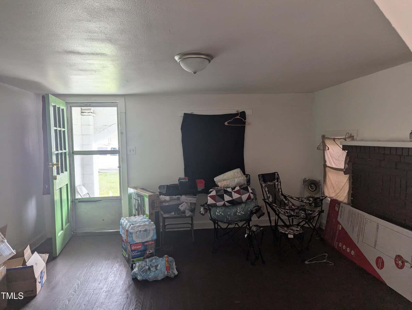 A small room with minimal furniture. The room has a green front door, a window covered with a black cloth, and a light fixture on the ceiling. The space contains folding chairs, assorted items including bottled water, and a few boxes on the floor.