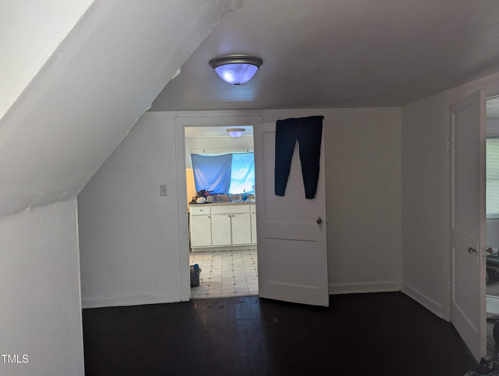 A small, unfurnished room with dark wooden floors, white walls, and a partially open door leading to another room with a blue-tinted window covering. A pair of jeans hangs over the door. Ceiling lights are on in both rooms.