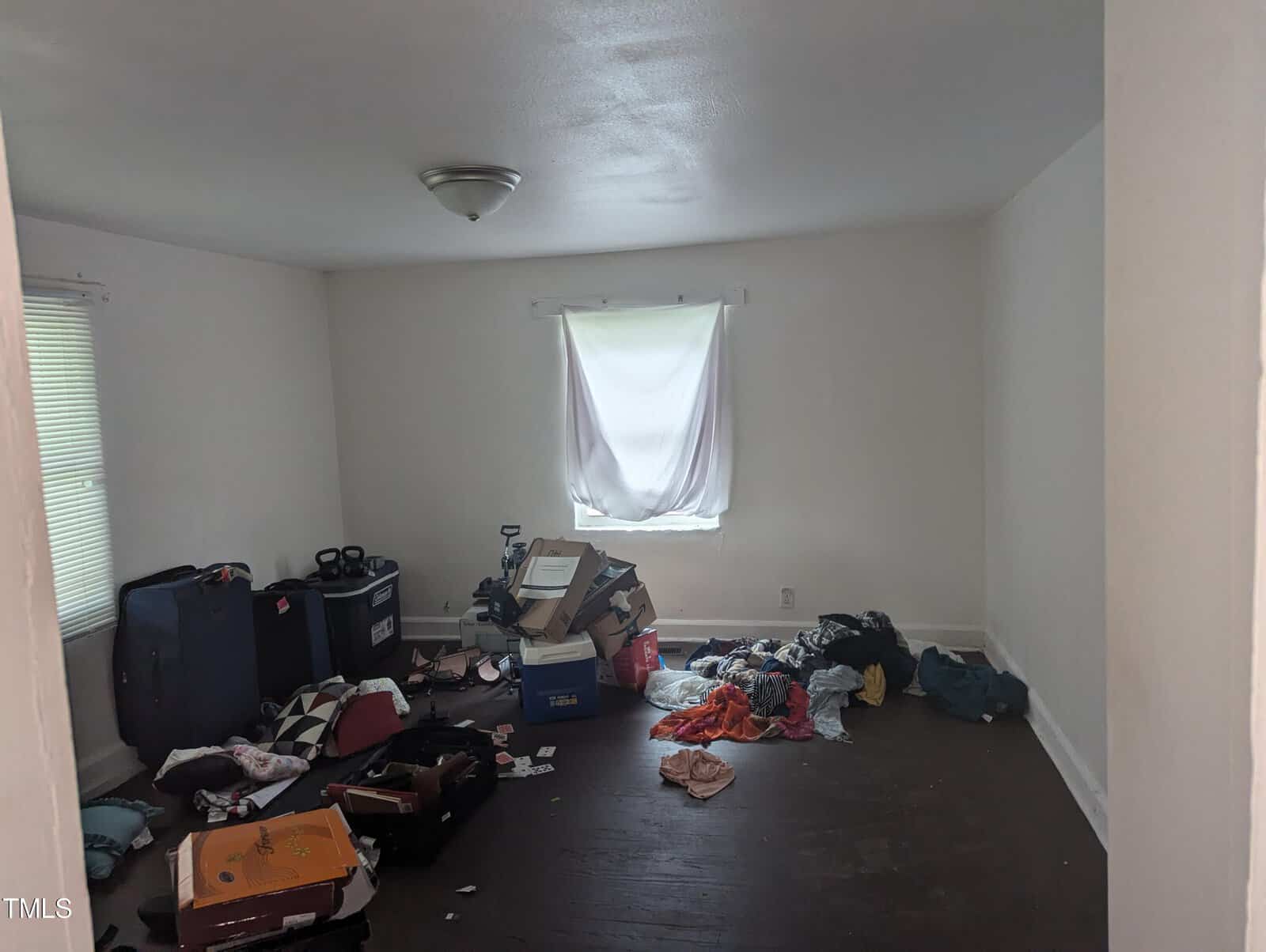 A room with white walls and dark flooring, containing scattered clothes, boxes, bags, and a suitcase. A white sheet covers the window. The room appears cluttered and dimly lit.