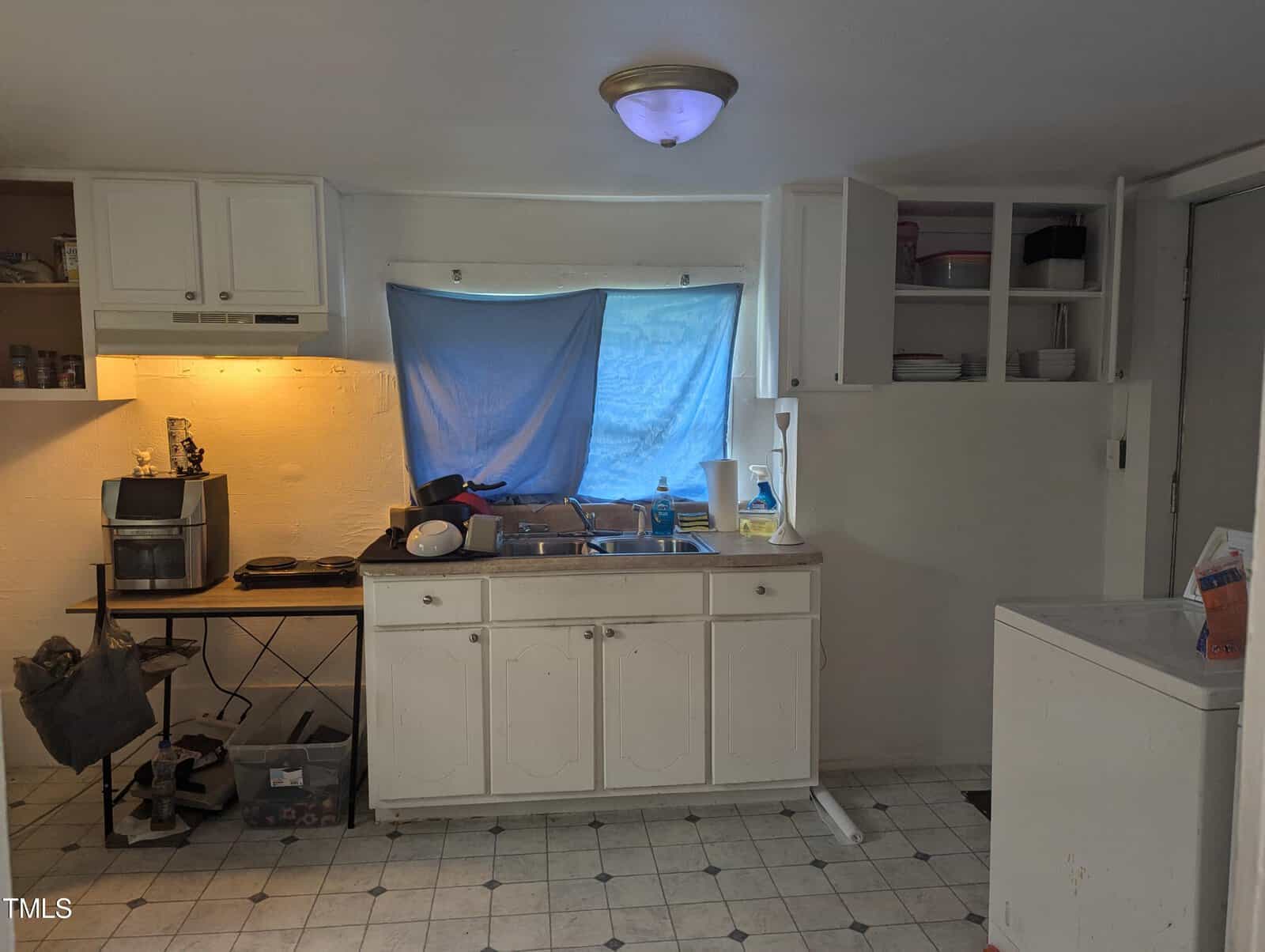 A small kitchen with white cabinets and appliances. A blue cloth covers the window. The countertop holds various household items, and there is a coffee maker on a small table. Some shelves are open, showing dishes. The floor has a light-colored tile pattern.