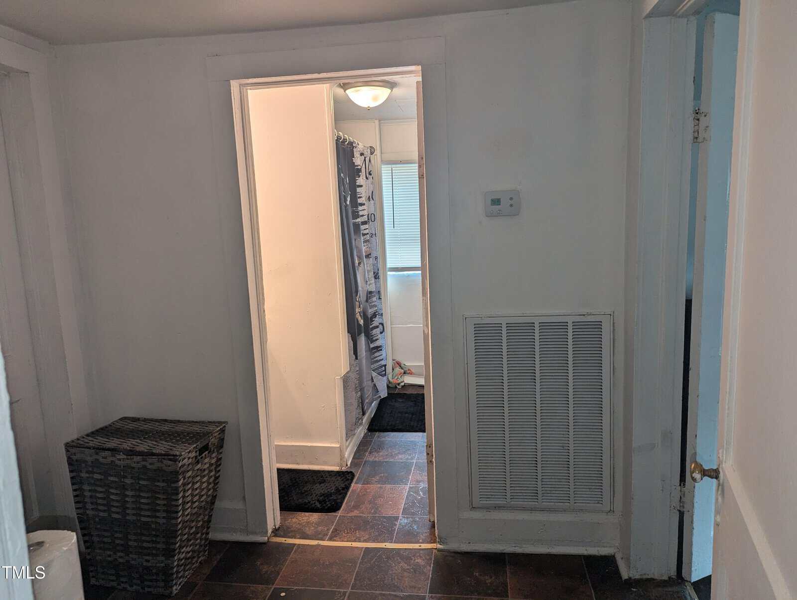 A small hallway in a home, featuring a door leading into a bathroom with a tiled floor. The bathroom has a shower with a curtain, and a window with blinds. To the left, there is a woven laundry basket. Walls and doors are painted white.