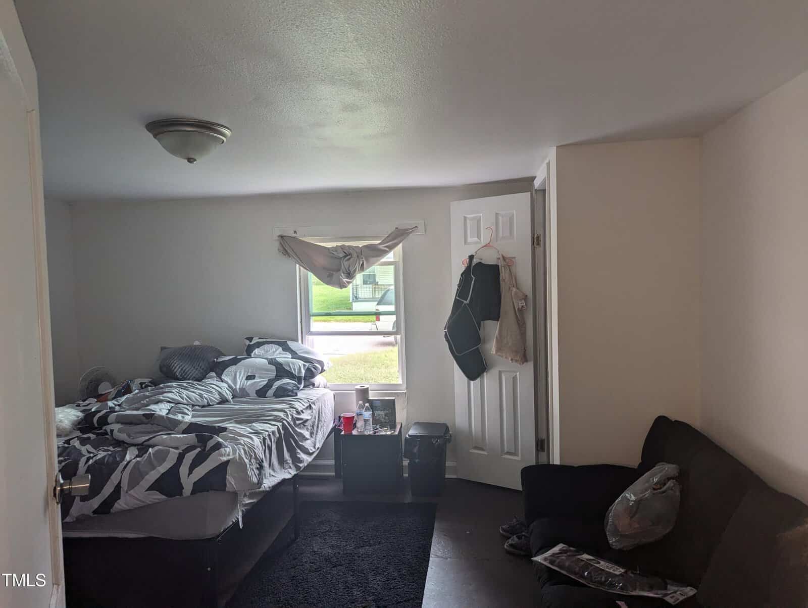 A small bedroom with a bed on the left, covered in a disheveled black, white, and gray patterned comforter. There is a window with a wrinkled drape. On the right, a black couch sits near an open door with bags and jackets hanging on it. The floor is carpeted.