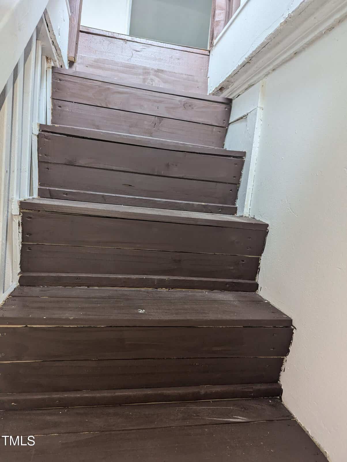 A set of dark wooden stairs with white walls on either side, leading up to a landing. The wooden steps have visible wear and tear. A white banister is partially visible on the left side of the stairs.