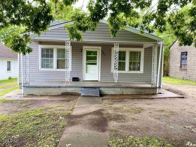 A small single-story house at 327 Hamilton Street, Henderson, with gray siding and a white front door is pictured. It features a covered porch with decorative metal railings and two large windows flanking the door. The surrounding yard is grassy, with some trees providing shade.