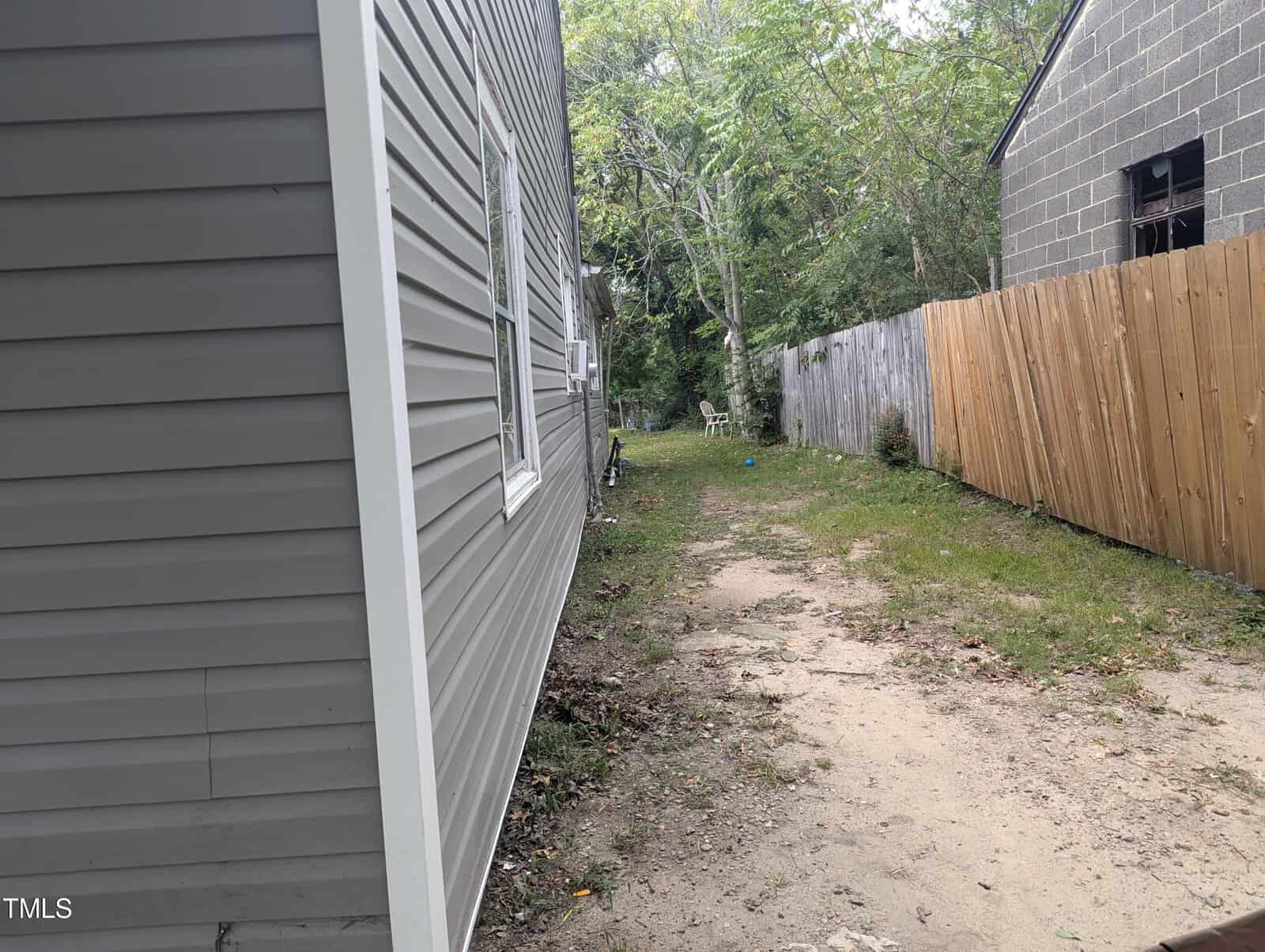 A narrow, grassy pathway runs between two houses. The house on the left has gray siding and a white framed window, while the house on the right has gray concrete blocks and a wooden fence. In the distance, some trees and a white chair are visible.