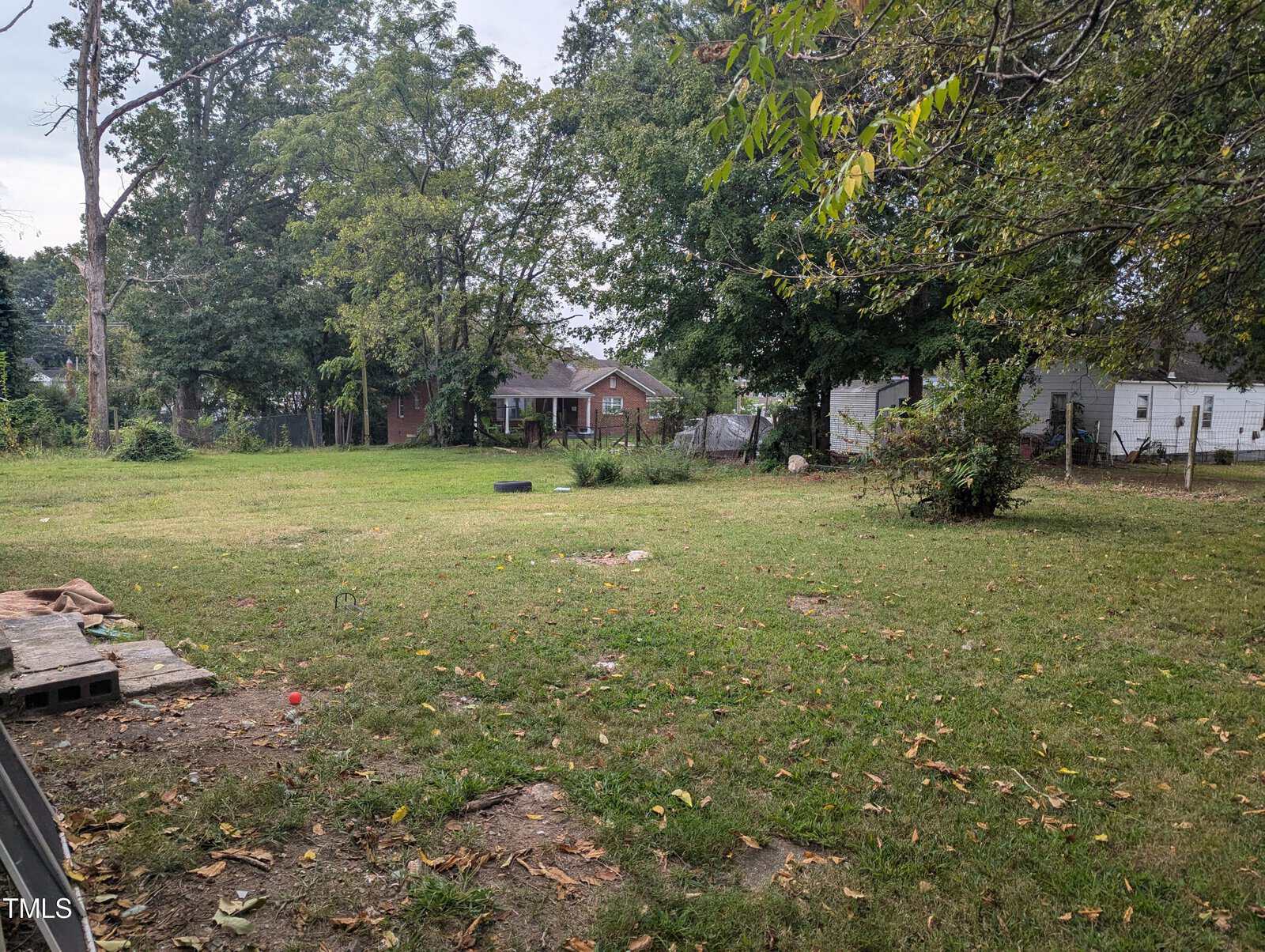 A spacious, slightly overgrown backyard surrounded by trees with a red-brick house partially visible in the background. The area features scattered debris, including wooden pallets and tires, and a small white shed on the right side.