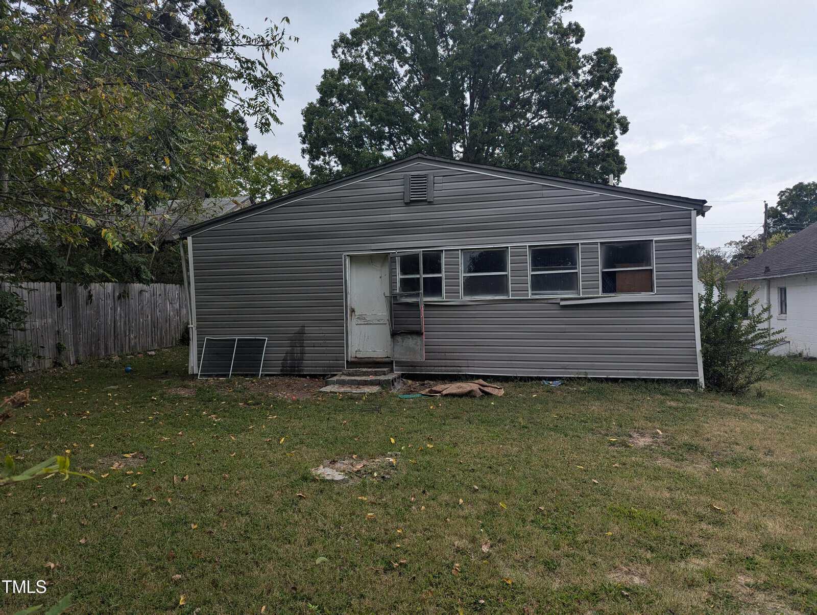 A slightly weathered, single-story house with grey vinyl siding and a small, elevated porch. The property has a fenced-in backyard with patches of grass and some scattered debris. Several windows are visible, with one partially boarded up. Trees are in the background.