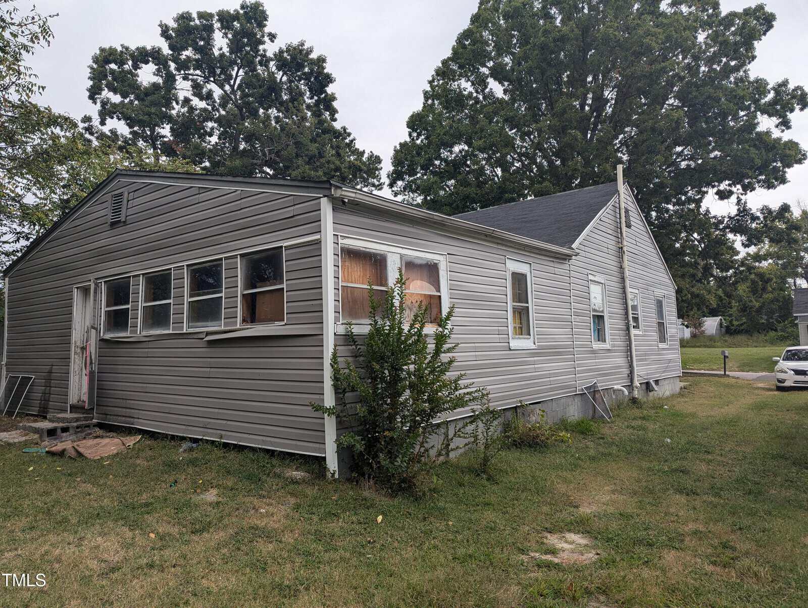 A one-story house with light gray vinyl siding has several windows, some of which are covered. A tall bush grows near the side of the house. The lawn is patchy with grass and dirt. A white car is parked in the background near a large tree.