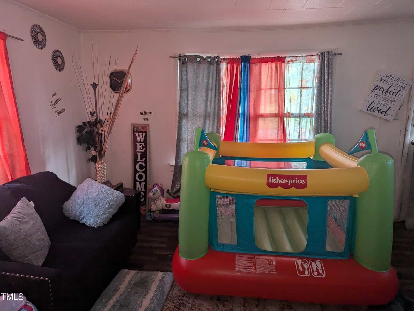 A cozy living room with colorful curtains and a children's inflatable bounce house from Fisher-Price beside a dark-colored sofa. The walls have decorative items, including a tall plant, a "Welcome" sign, and motivational quotes. The floor has patterned rugs.