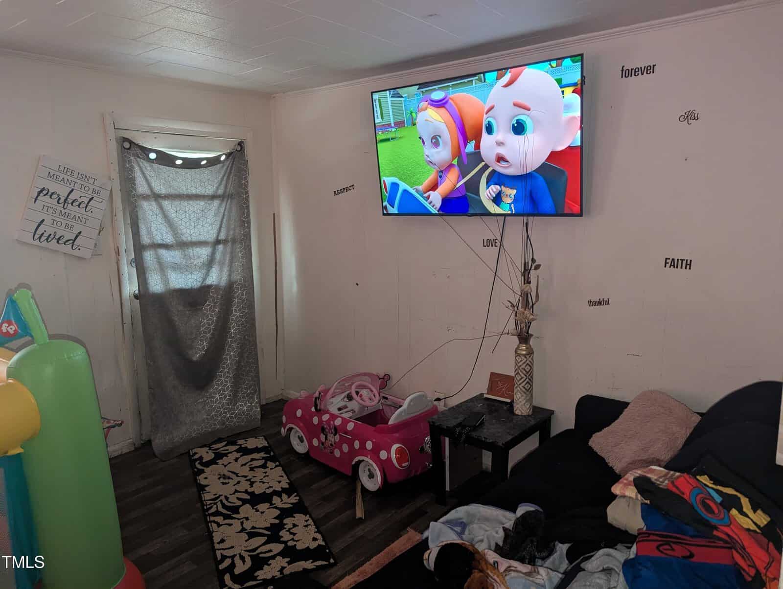 A cluttered living room with a cartoon playing on a wall-mounted TV. Toys are scattered, including a small pink toy car. Inspirational wall decals and a black couch with blankets are visible. A curtain covers the entrance.