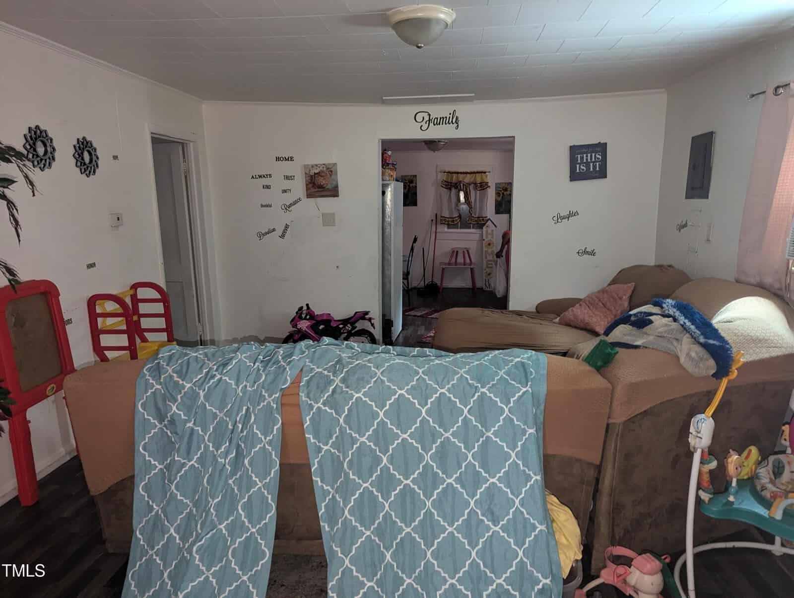 A cozy living room with a couch covered in a blue patterned blanket and several children's toys scattered around. The walls are decorated with family-themed quotes. A window allows natural light to brighten the space.