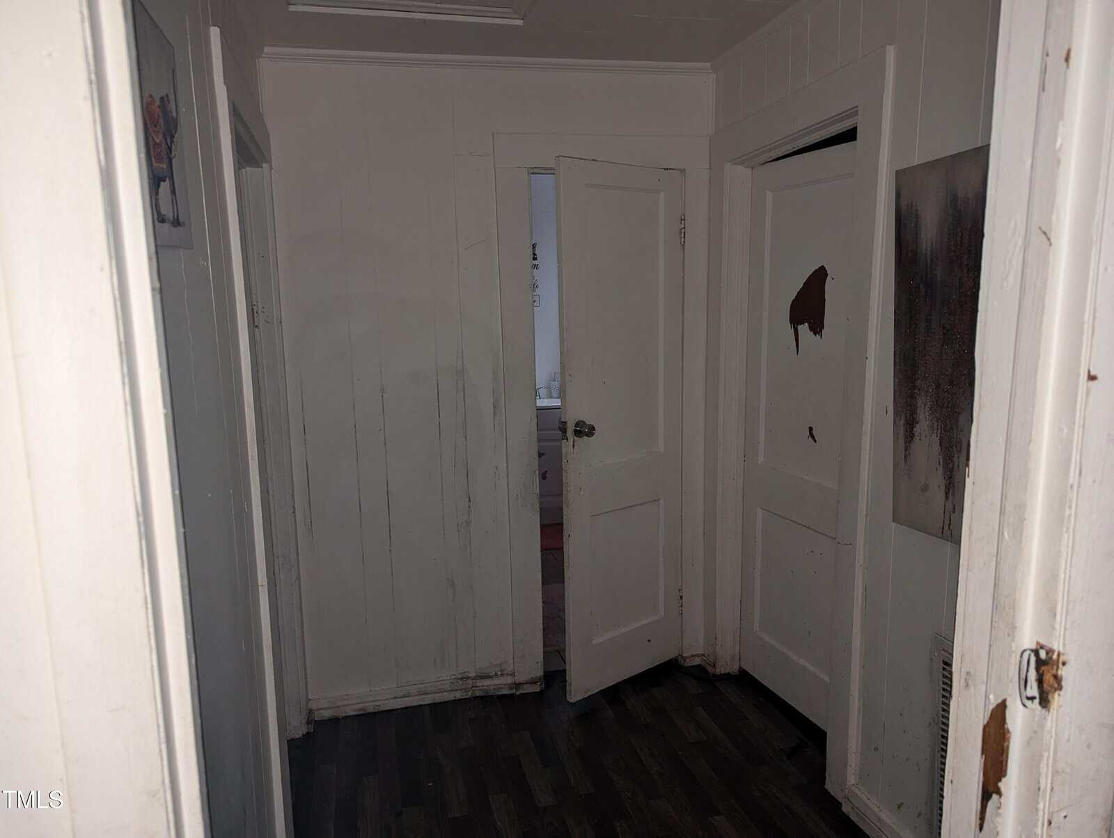A dimly lit, narrow hallway with white paneled walls and dark wooden flooring. Three doors are visible: the closest on the left is partially open, the middle door is ajar, revealing a glimpse of another room, and the right door is closed with visible damage.