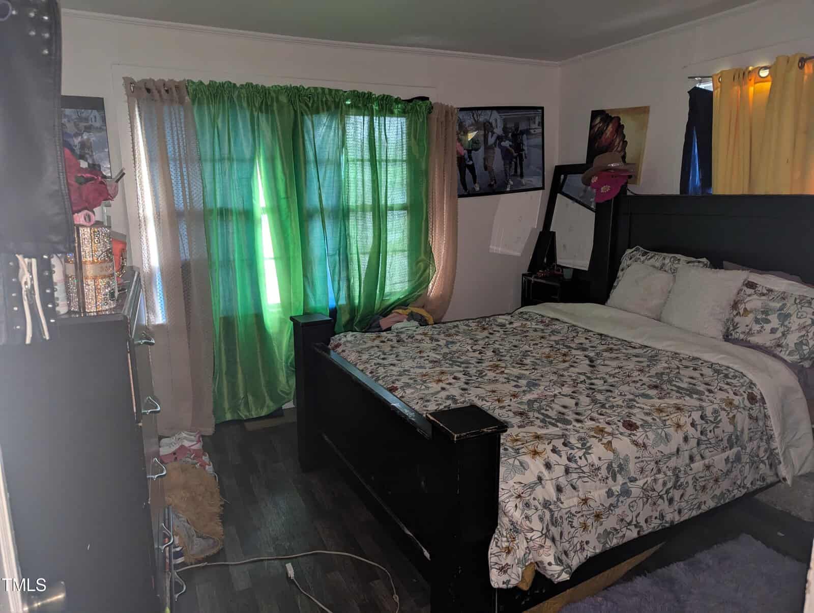 A bedroom with dark wood flooring and a bed with a floral comforter. Two windows are covered with green and gray curtains. The room contains a cluttered shelving unit, a framed picture on the wall, and various items scattered on the floor and furniture.