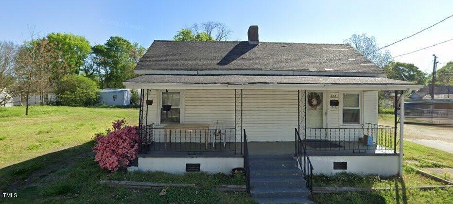 A single-story white house at 326 Hamilton Street in Henderson features a front porch with steps and a metal railing. The porch has a table, a chair, and a wreath on the door. A small pink bush sits to the left of the stairs, surrounded by green grass in the yard. Trees and open space are visible in the background.