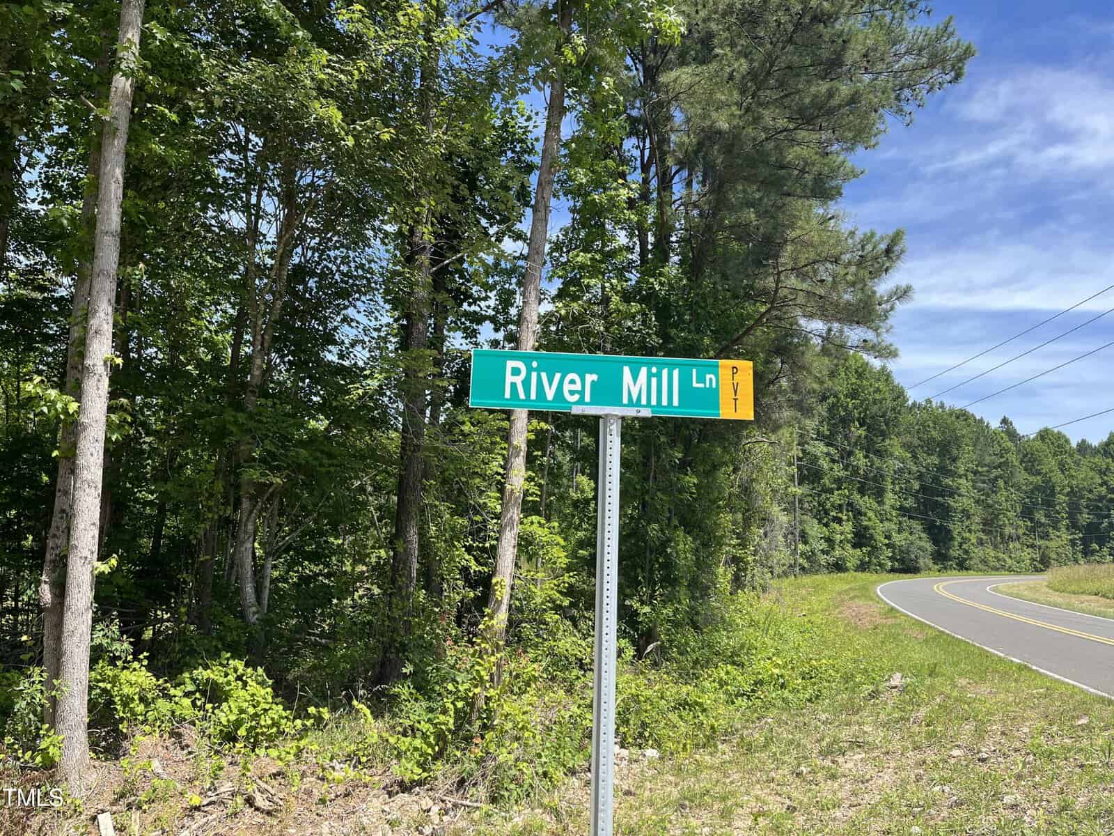 Street sign for River Mill Ln at a forested intersection with a winding road. Tall trees and a clear sky are visible in the background.