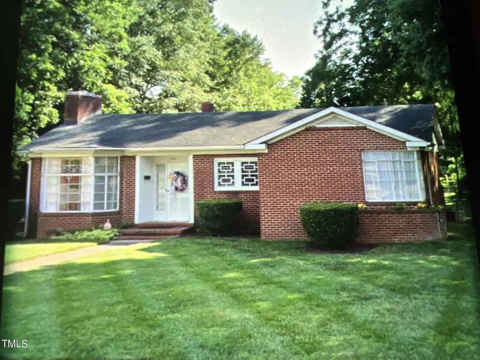 A single-story red brick house with a dark roof and white-framed windows graces 203 W Rockspring Street in Henderson. The white front door, adorned with a wreath, is flanked by bushes. The yard is impeccably maintained, and stately trees provide a picturesque backdrop.