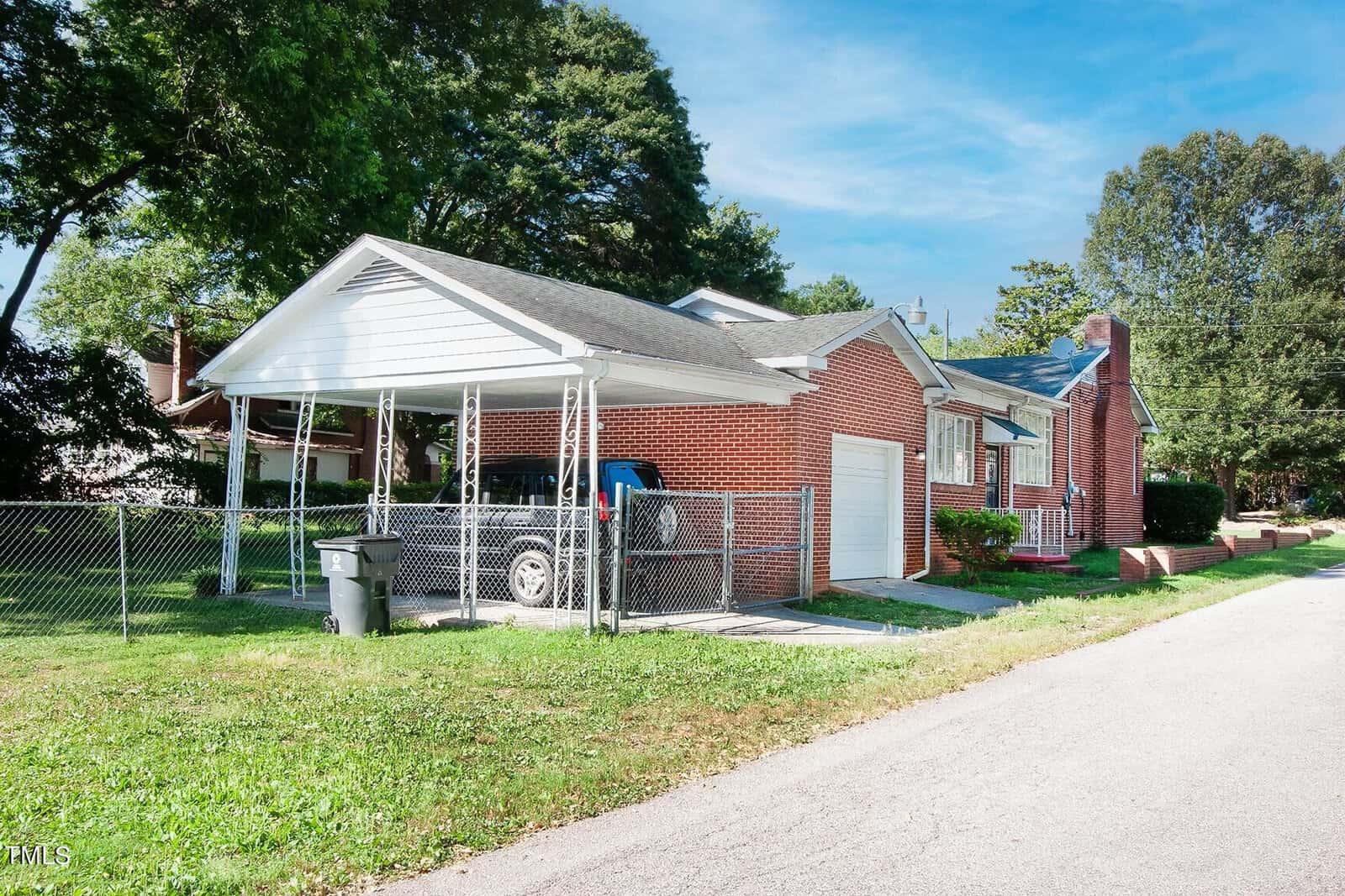 A suburban brick house with a carport is seen, featuring a fenced yard and a paved driveway. The carport contains a dark-colored vehicle. Green trees and a clear blue sky form the backdrop, and a paved road runs alongside the property.