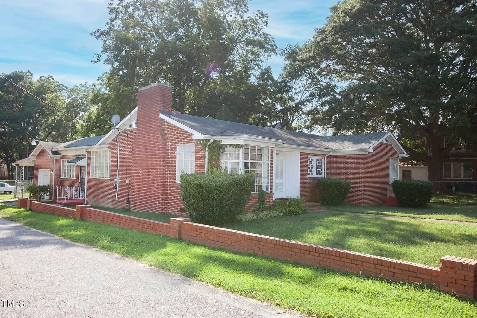 A red brick, single-story house with a chimney, large front windows, and a manicured lawn. There are bushes in front and a low brick wall bordering the yard. The house is situated on a corner lot with trees in the background under a clear, blue sky.