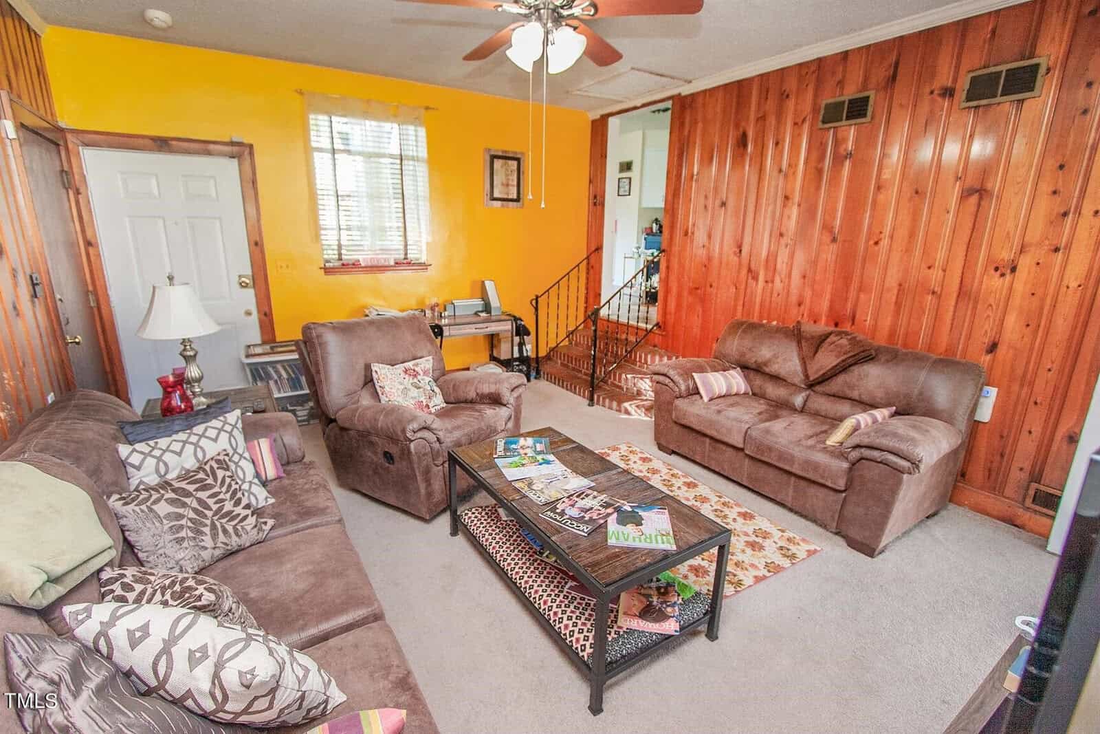A cozy living room with wood-paneled walls and a carpeted floor. It has two brown sofas, a central coffee table with magazines, a floor lamp, a ceiling fan, and a desk with a printer near the back wall. A staircase leads to another area of the home.