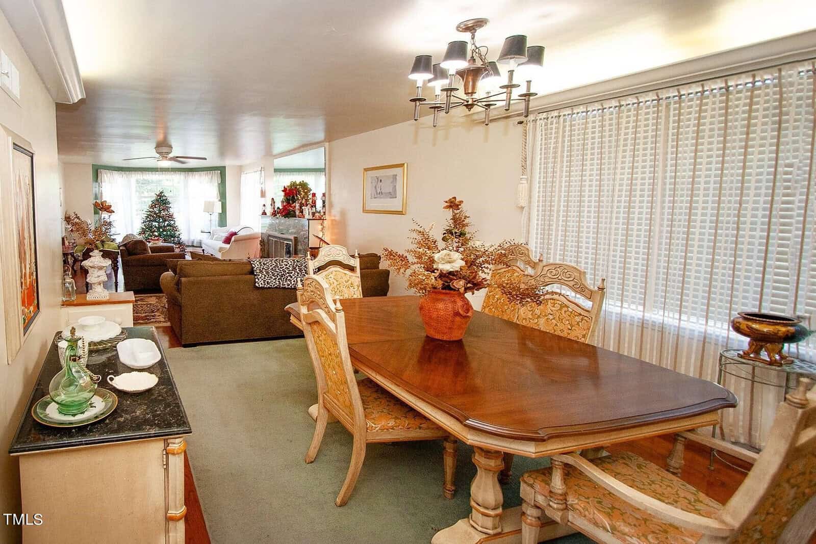 A dining area with a wooden table and six upholstered chairs leads into a cozy living room. Both spaces are decorated with various plants and a Christmas tree. A chandelier hangs above the dining table, and large windows with vertical blinds allow light in.
