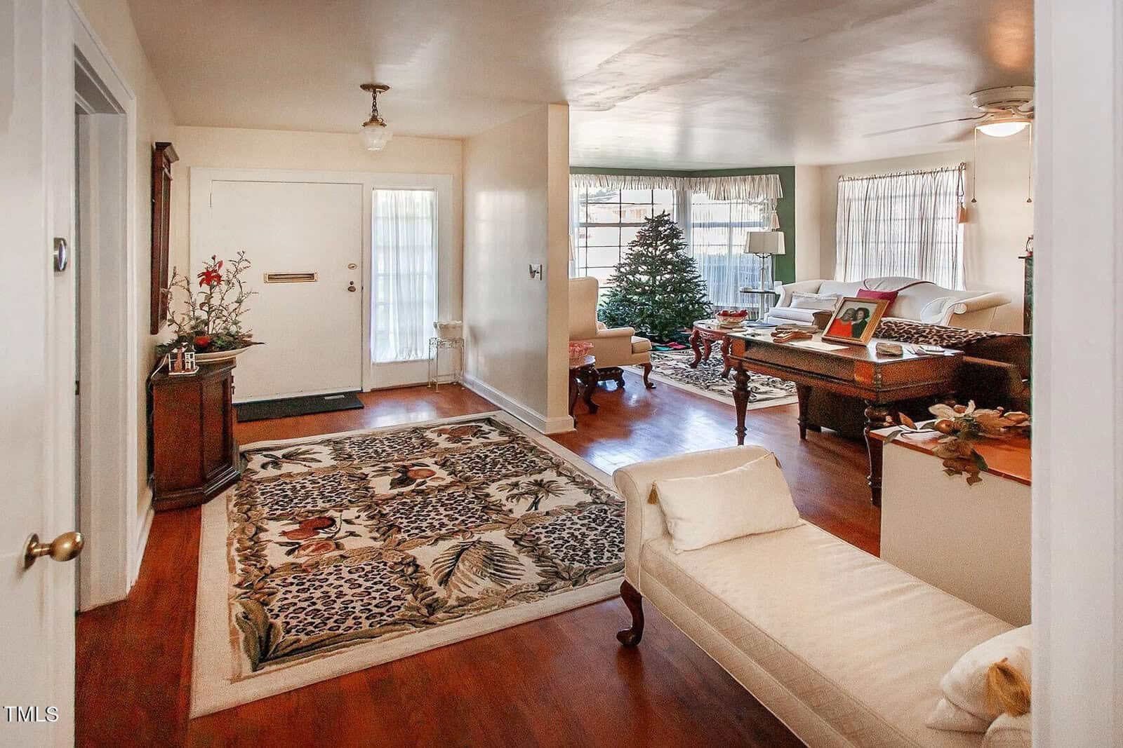 A cozy living room features a decorated Christmas tree by large windows. The room has wooden floors, various upholstered seating, a white chaise lounge, a patterned area rug, and a front door with a small chandelier above. Light filters in through sheer curtains.
