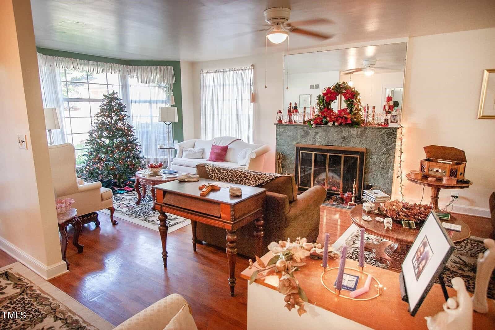 A cozy living room decorated for Christmas, featuring a Christmas tree by the window, a fireplace adorned with holiday decorations and a wreath, several sofas and armchairs, and wooden furniture. The room is warmly lit with natural light streaming through sheer curtains.
