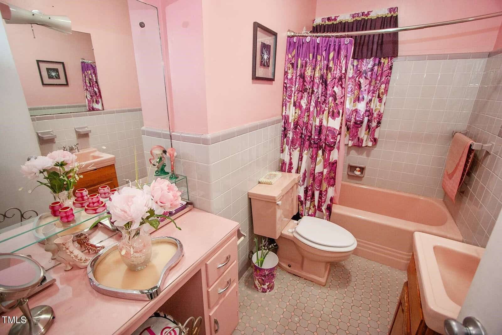 A retro-style bathroom with pink walls and a pink bathtub. There is a floral shower curtain, a toilet, and a sink with a pink countertop. The sink area has decorative items such as flowers, a tray, and toiletries. A framed picture hangs on the wall.