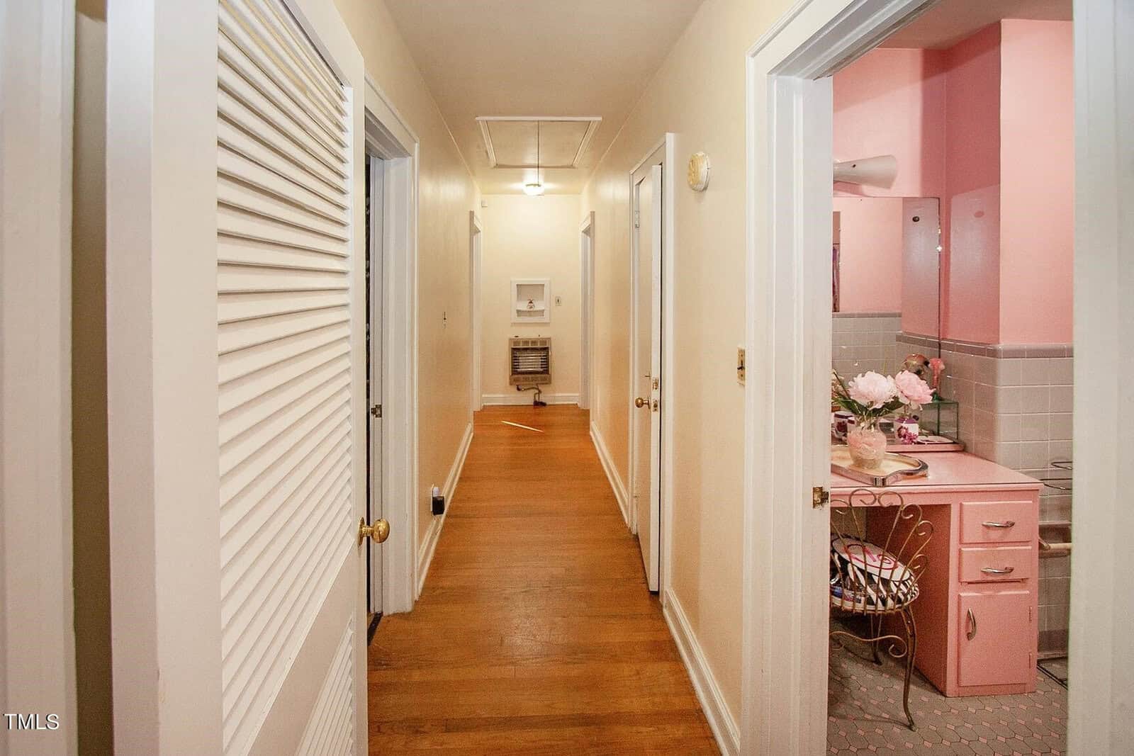 A hallway with wooden flooring and cream-colored walls. There are several white doors along the hallway. On the right, an open door reveals a retro pink-themed bathroom with a pink sink, vanity, and checkered flooring. A closet with louvered doors is on the left.
