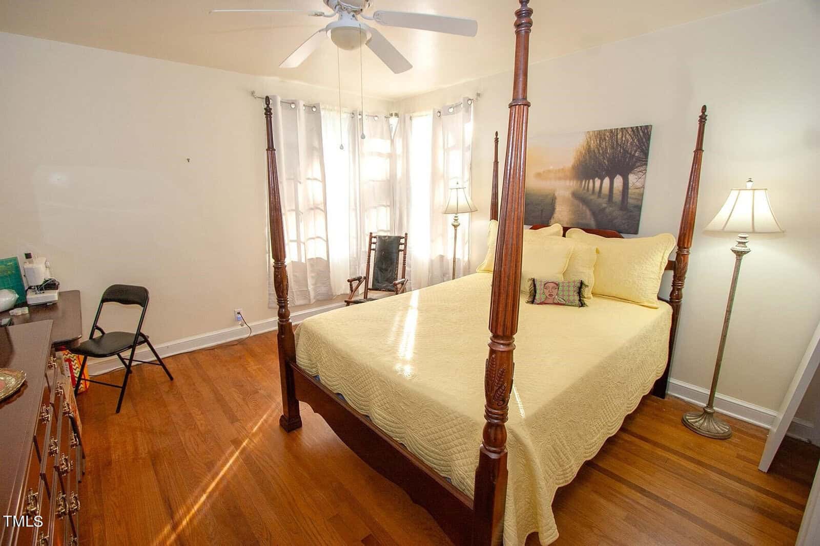 A bright bedroom with a polished wooden floor features a four-poster bed covered with a yellow quilt. The room has a ceiling fan, a tall floor lamp, a rocking chair near a window with sheer white curtains, and a dresser with a folding chair beside it.