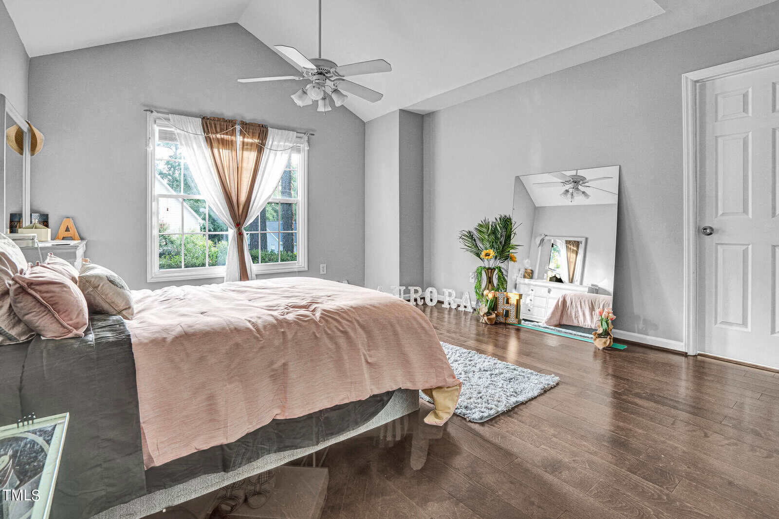 A cozy bedroom with a large bed covered in pink and green bedding, situated near a window with sheer curtains. There's a floor mirror reflecting a potted plant, and a light blue rug on the wooden floor. Ceiling fan overhead and soft natural light entering.