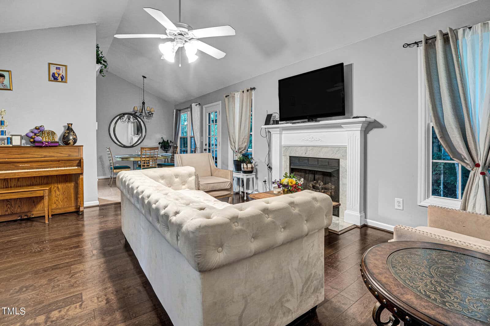 A cozy living room with a white tufted sofa facing a wall-mounted TV above a fireplace. A ceiling fan hangs above. A piano is on the left, and a dining area with a large mirror is visible in the background. Light curtains frame the windows.