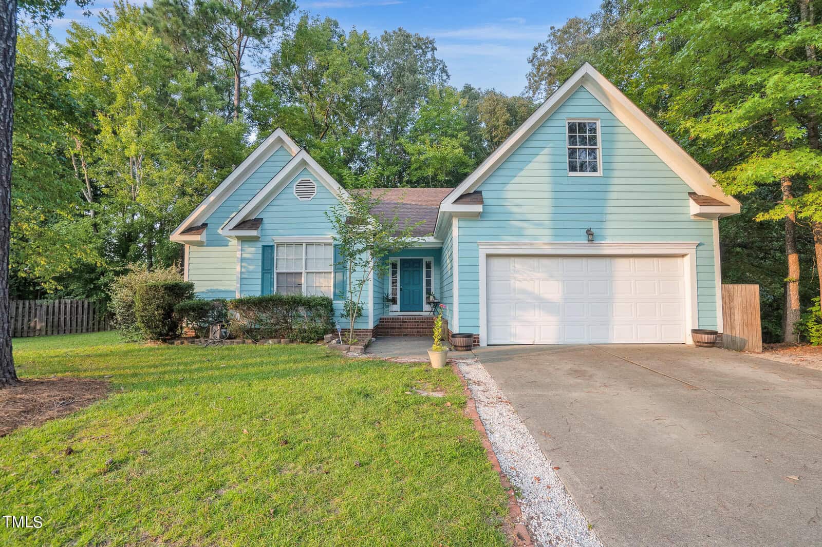 Nestled in a wooded suburban area, this charming light blue house at 2745 Michelle Ct, Creedmoor, features a pitched roof and an attached two-car garage. The driveway unfurls gracefully from the garage, while a quaint walkway welcomes you to the entrance amidst lush green trees.