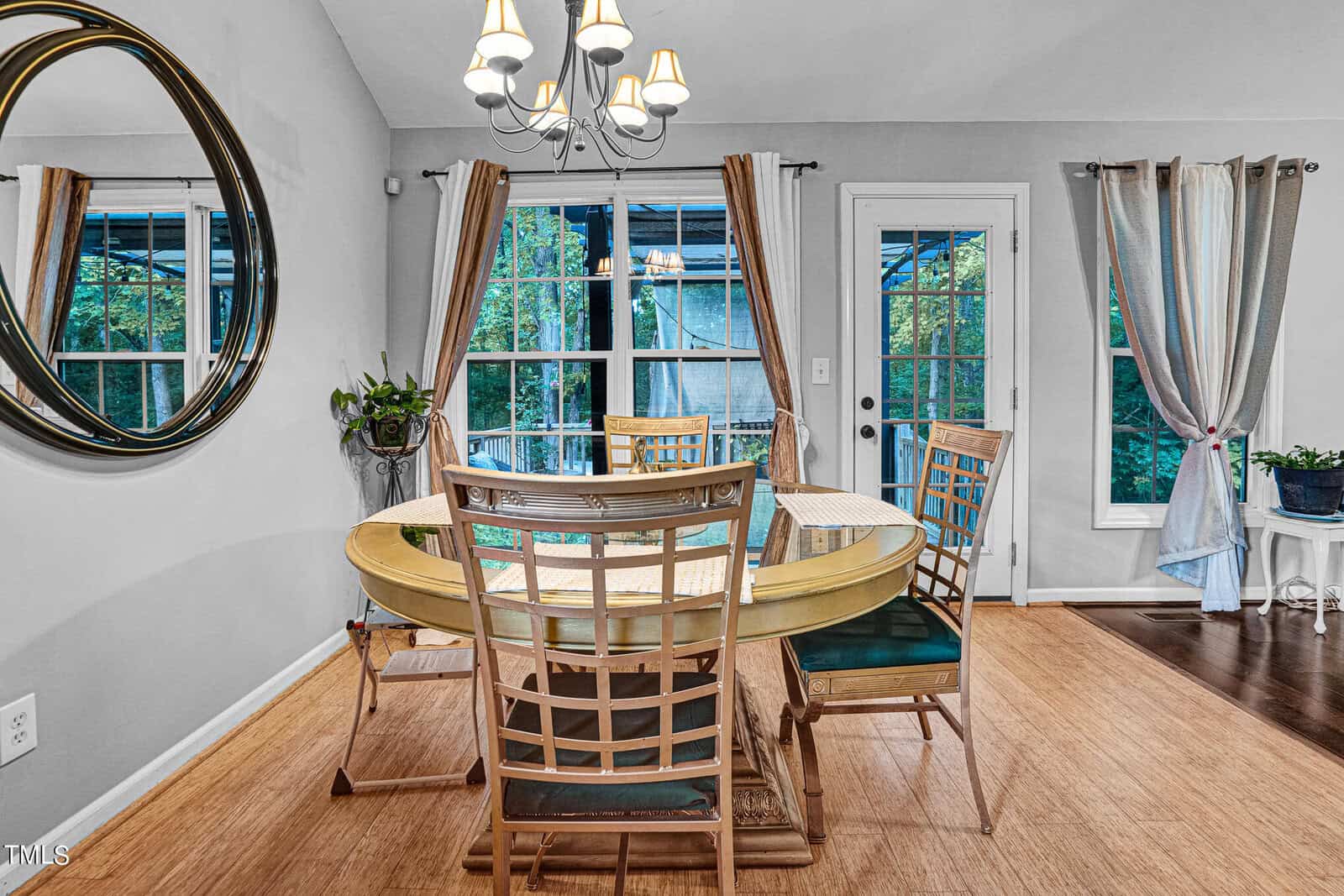 A dining room with a round wooden table and four chairs. A large circular mirror hangs on the wall. Windows with curtains bring in natural light. A potted plant sits on a side table, and a chandelier hangs above the table.