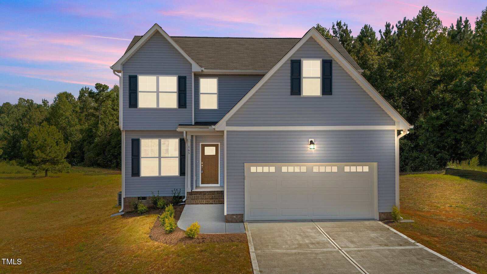 A two-story gray house with white trim and dark shutters, featuring a front porch, a double garage, concrete driveway, and a small landscaped area, set against trees and a sunset sky.