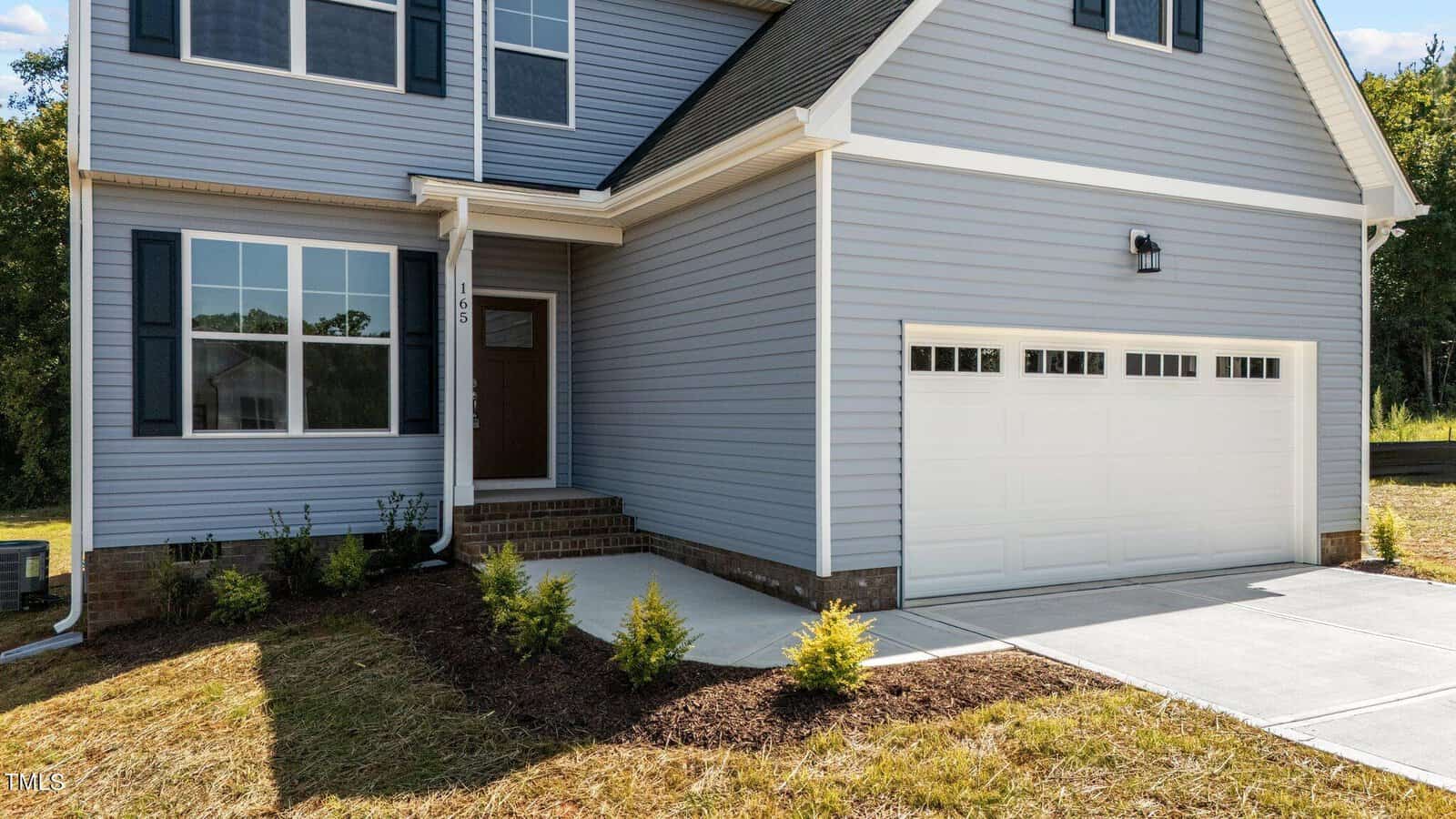 A two-story modern house with gray siding, white trim, a double garage door, and a brown front door. There are a few small shrubs and freshly laid mulch in the landscaped front yard.