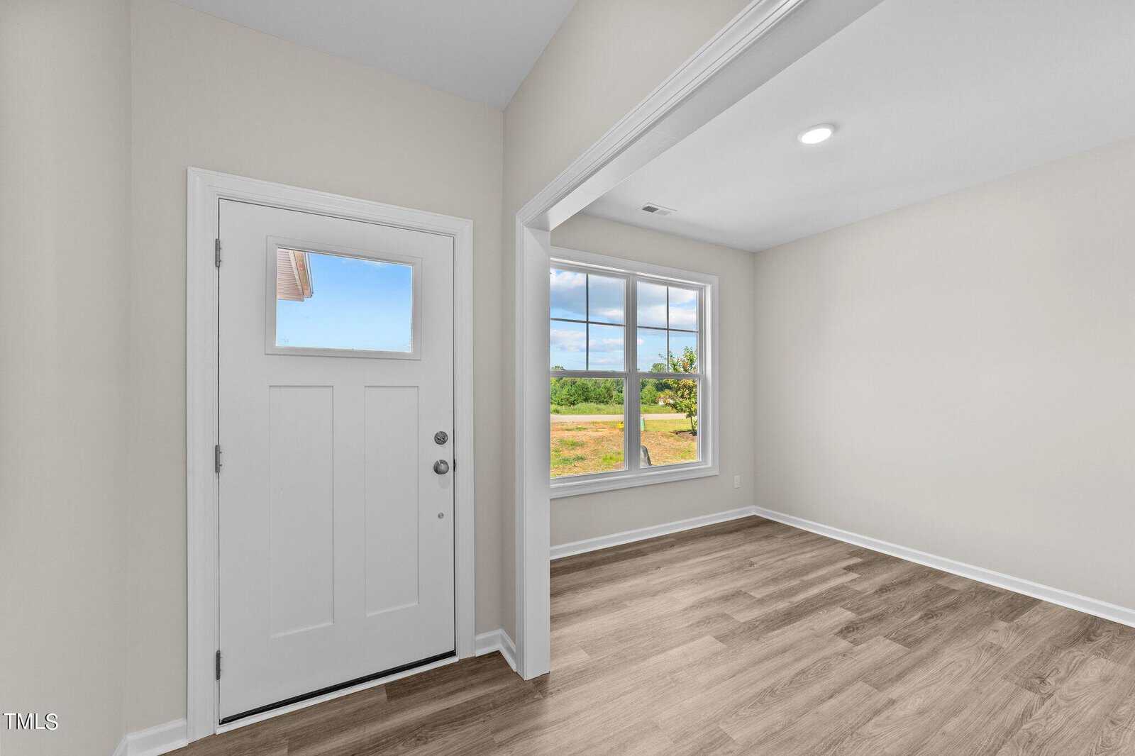 A bright entryway with a white front door on the left, large window with outdoor view on the right, and wood-style flooring throughout. Walls are painted light beige and the space appears uncluttered and modern.