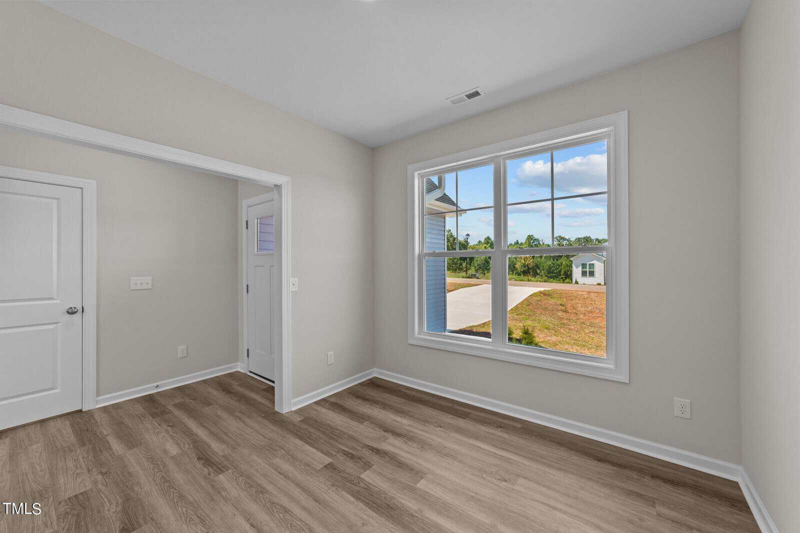 A small, empty room with light beige walls, wood-style flooring, a large window showing an outdoor scene, and a door leading to a hallway with another door and window visible.