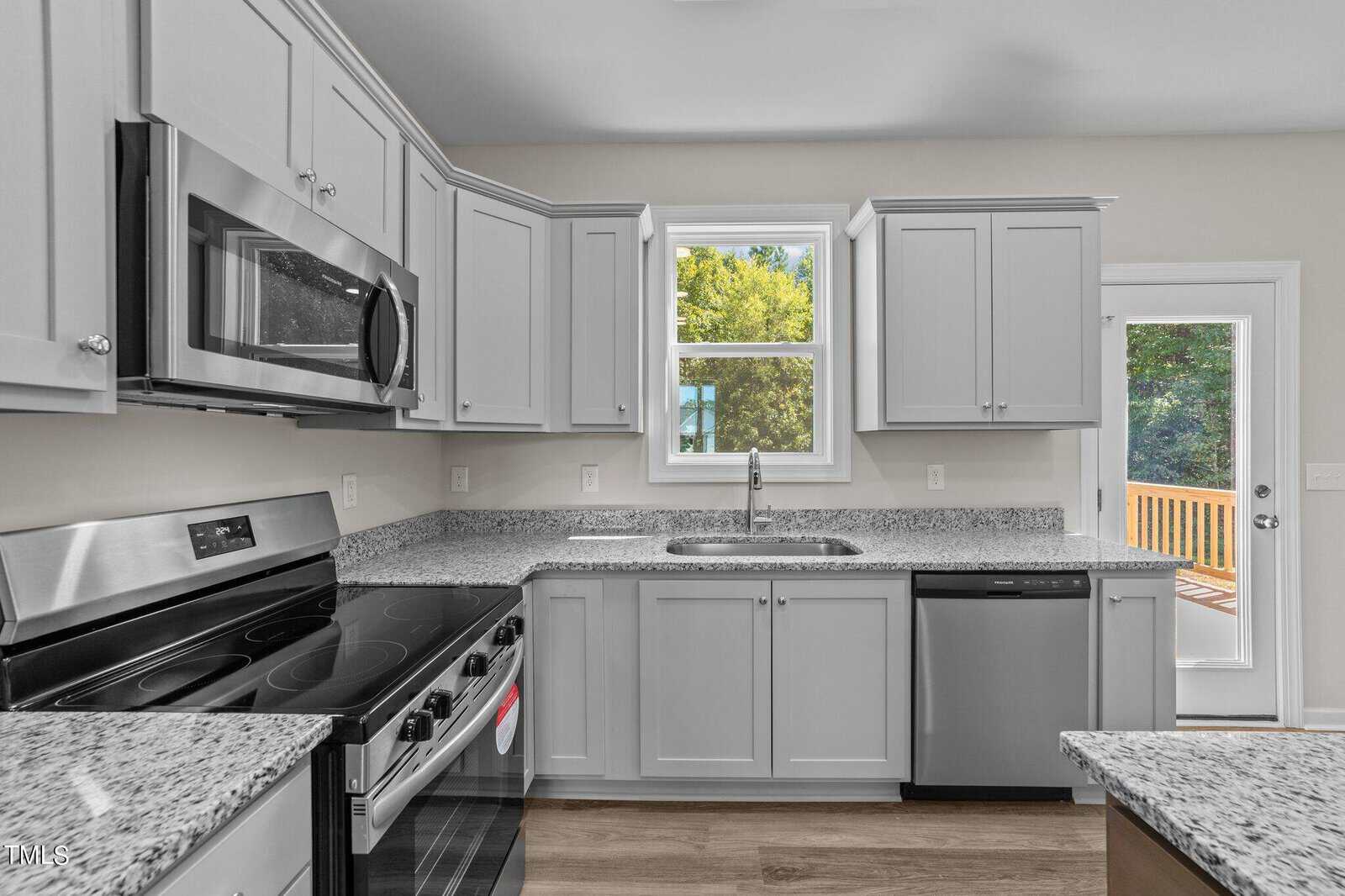 A modern kitchen with stainless steel appliances, light gray cabinets, granite countertops, a window above the sink, and a door leading outside. There is wood flooring and natural light coming in.