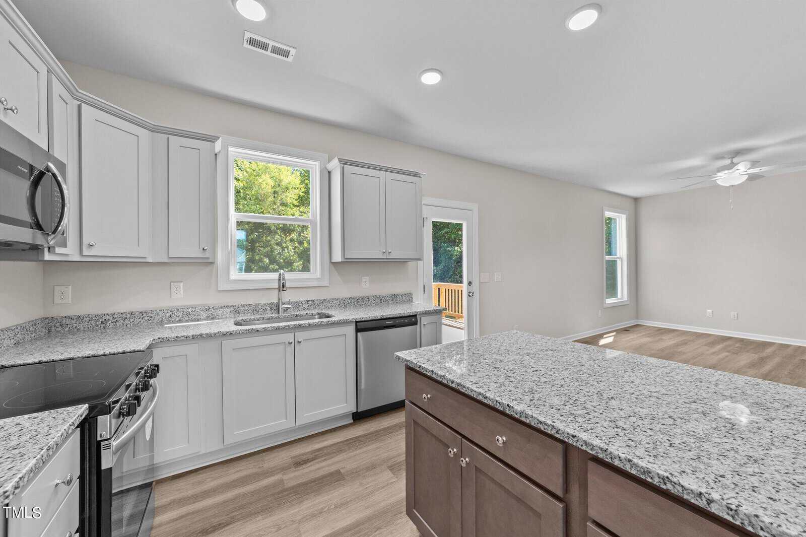 Modern kitchen with light gray cabinets, granite countertops, stainless steel appliances, and a large center island. The open layout connects to a living area with wood flooring and large windows. A door leads to an outdoor deck.