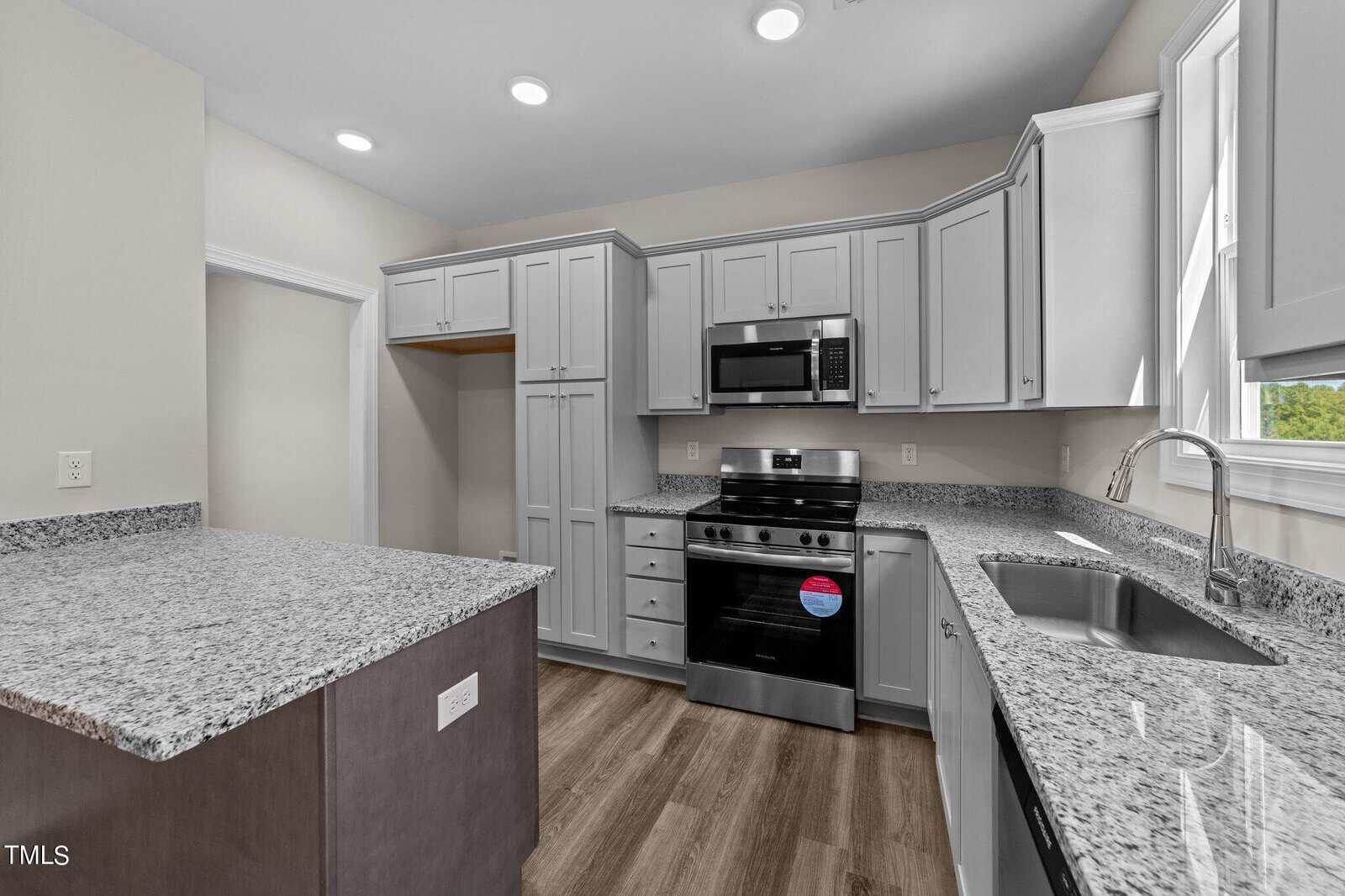 Modern kitchen with gray cabinets, stainless steel appliances, granite countertops, a double sink under a window, and wood-style flooring. A recessed area for a refrigerator is on the left. The space is well-lit with ceiling lights.