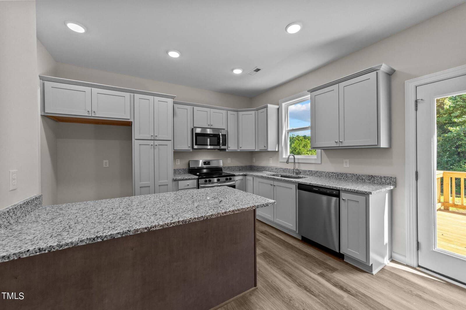 Modern kitchen with white cabinets, stainless steel appliances, granite countertops, a large island, wood flooring, and a glass door leading to an outdoor deck. A window provides a view of greenery outside.