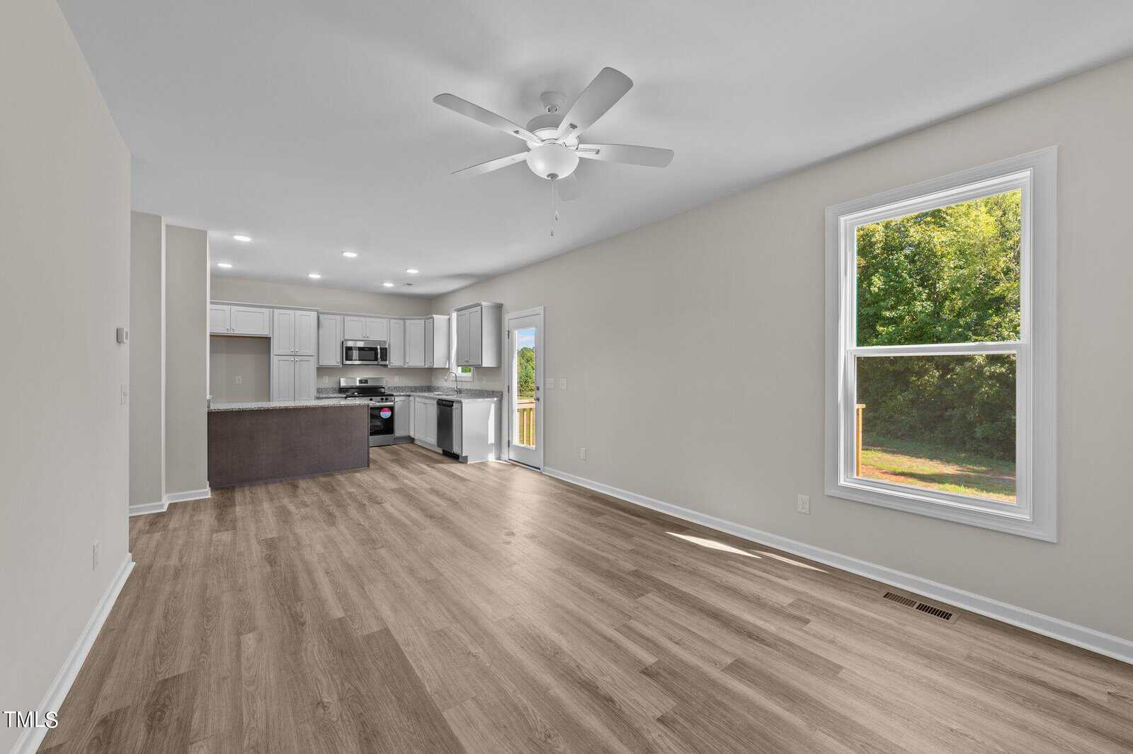 Open-concept kitchen and living area with light wood flooring, white walls, ceiling fan, and a large window overlooking greenery. The kitchen features white cabinets, stainless steel appliances, and recessed lighting.