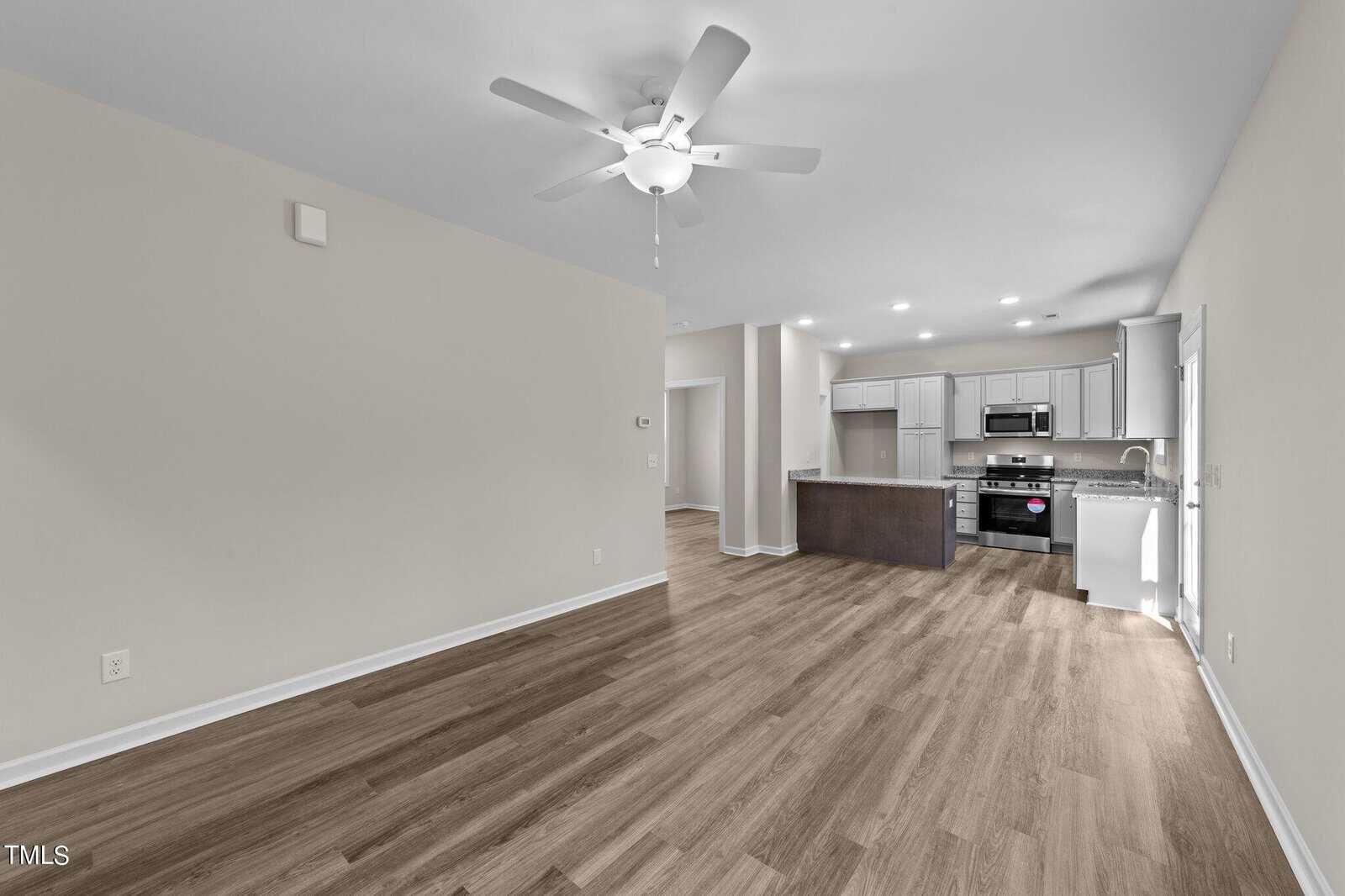 Empty modern living area with light wood flooring, beige walls, a ceiling fan, and an open kitchen with stainless steel appliances, white cabinets, and a center island. Multiple ceiling lights illuminate the space.
