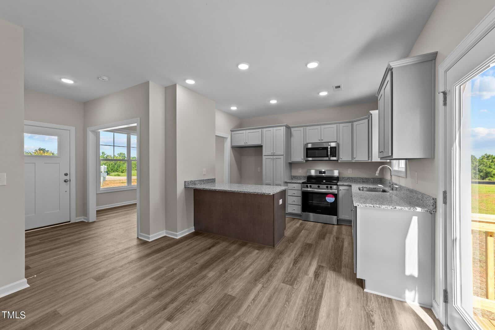 A modern kitchen with light gray cabinets, stainless steel appliances, granite countertops, and an island. The room has wood-style flooring, recessed lighting, and large windows providing natural light.