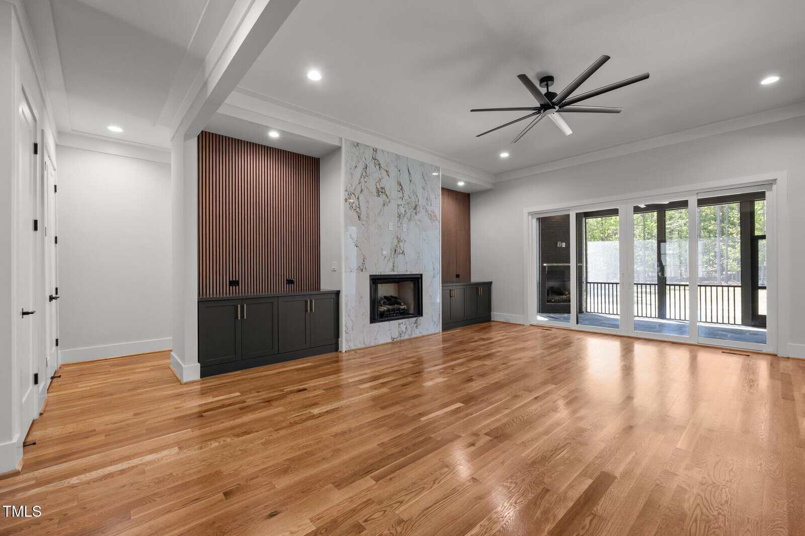 A spacious, modern living room with light wooden floors, a marble fireplace, dark built-in cabinets with vertical wood paneling, a ceiling fan, and large glass doors leading to a patio.