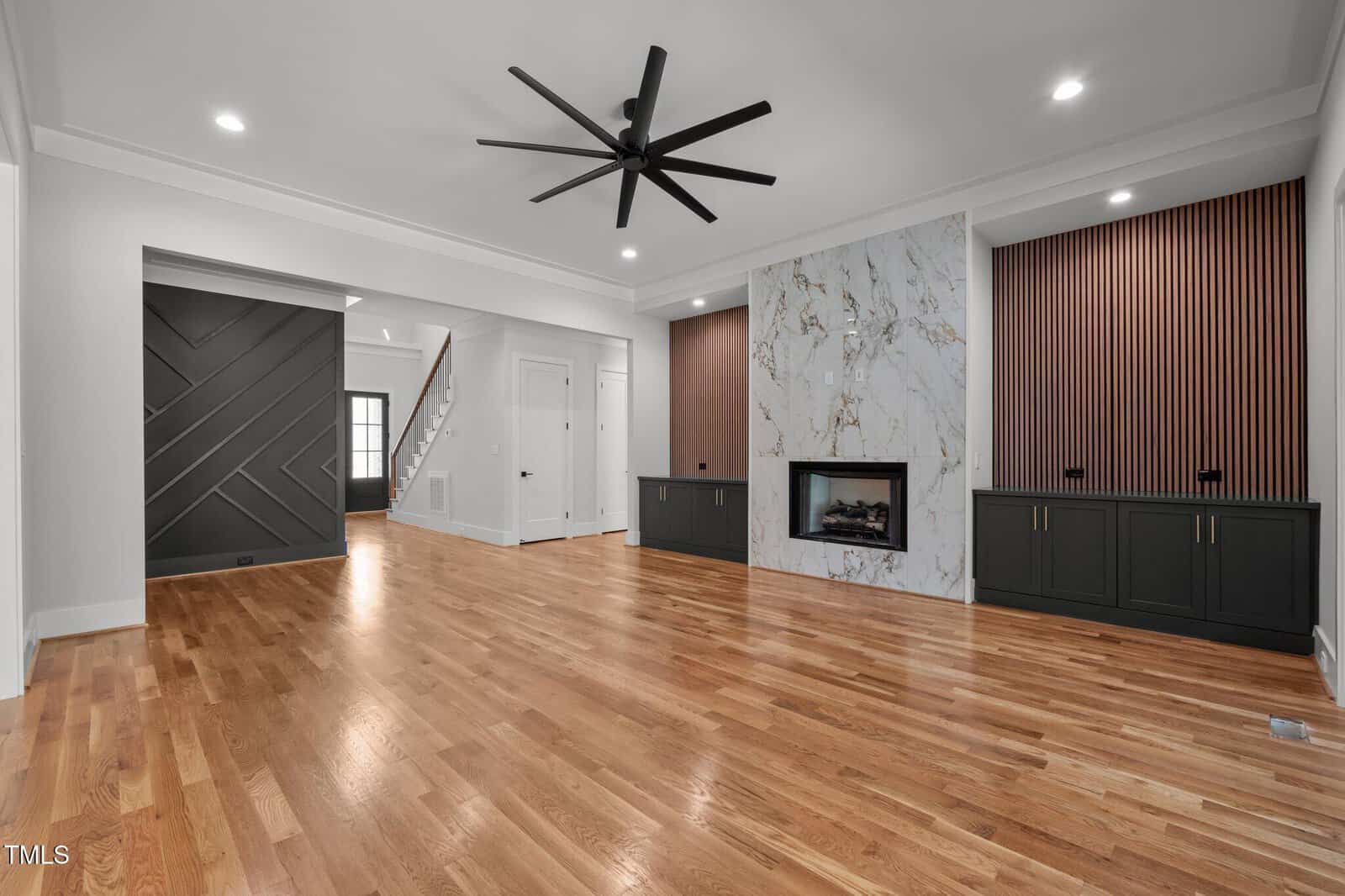 Modern living room with light wood floors, a marble fireplace, black built-in cabinets, slatted wood panels, a large black ceiling fan, and white walls with black geometric accent wall near a staircase.