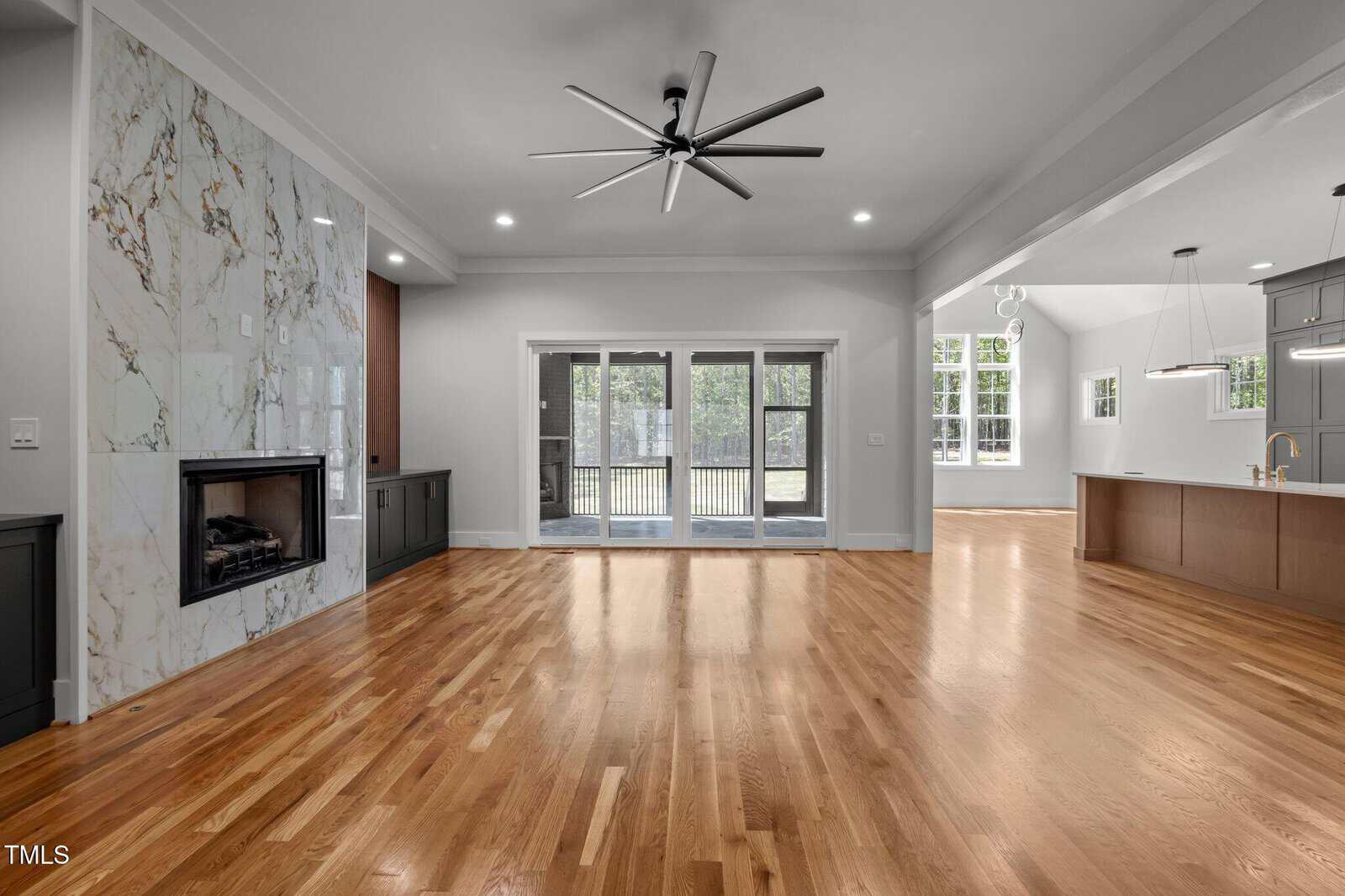 A spacious, modern living area with hardwood floors, a marble fireplace, large sliding glass doors leading to a patio, and an open kitchen with gray cabinets, pendant lighting, and tall windows.