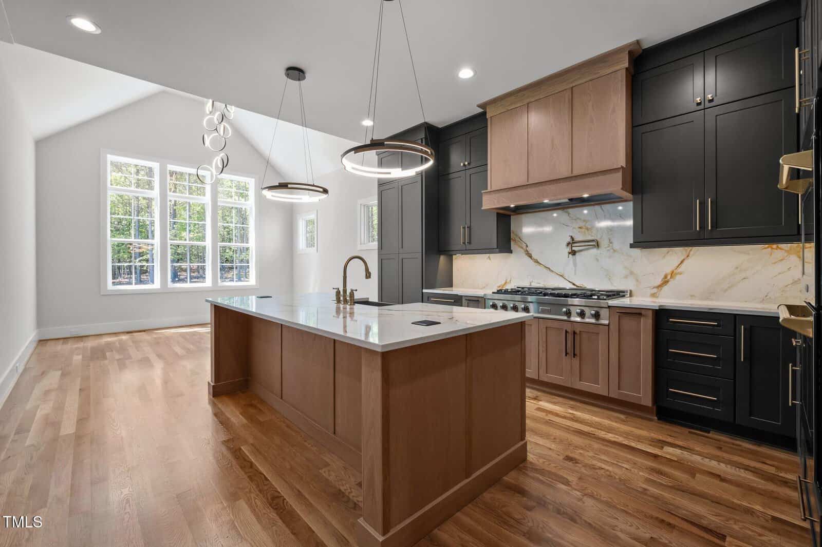 Modern kitchen with a large island featuring a wood base and white countertop, black and wood cabinetry, stainless steel appliances, marble backsplash, and two ring pendant lights. Hardwood floors and large windows provide natural light.