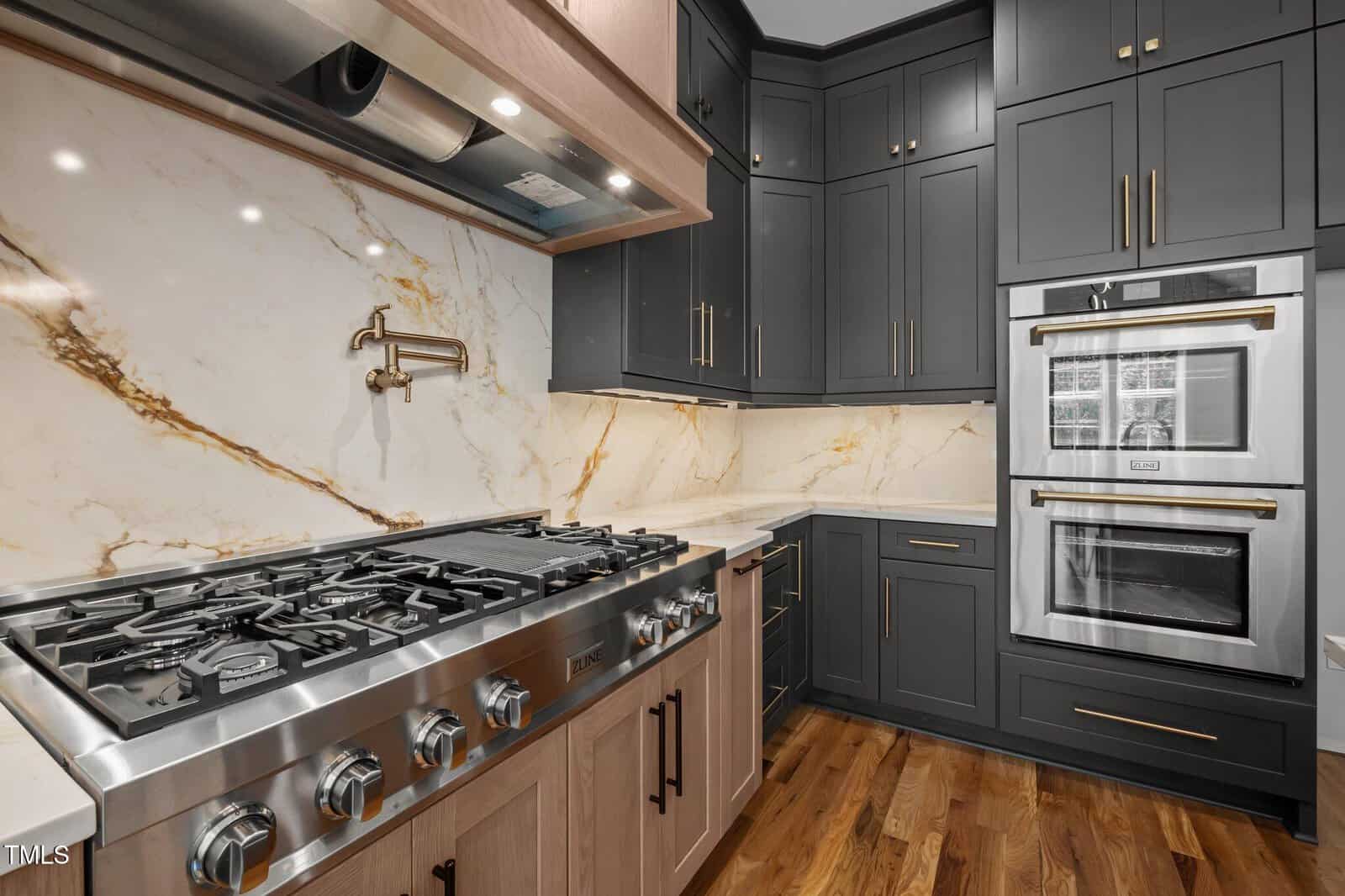 A modern kitchen with a stainless steel gas stove, marble backsplash with gold veining, black cabinets with gold handles, double wall ovens, and hardwood flooring.