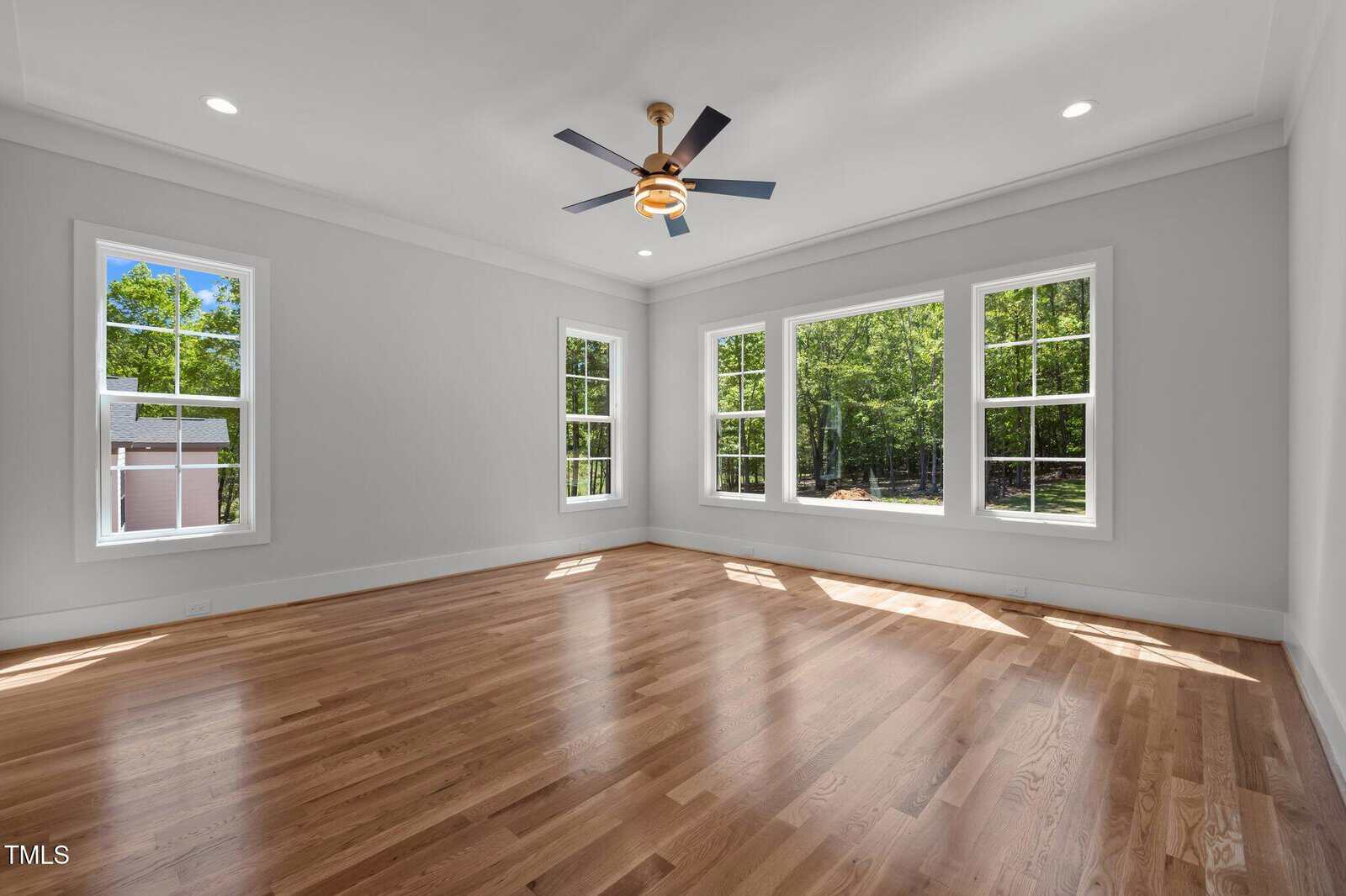 Bright, empty room with large windows, light gray walls, hardwood floors, white trim, and a ceiling fan with light. Sunlight streams in, highlighting the spacious, clean area. Trees and greenery visible outside.